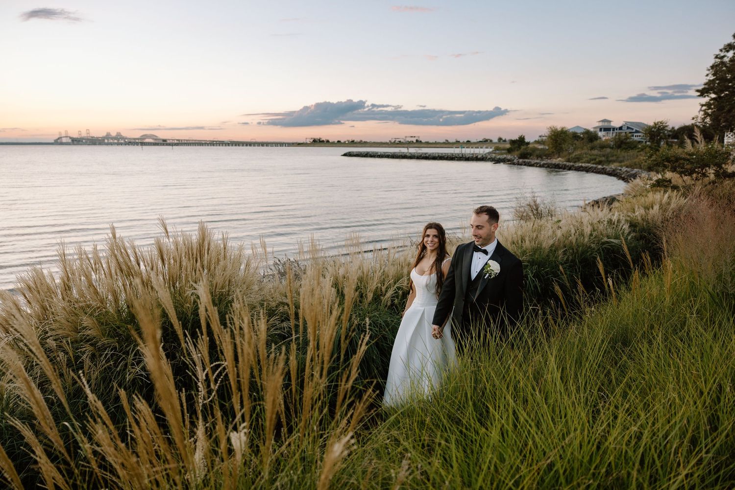 Bride and groom standing in tall grass by waterfront at sunset during romantic outdoor wedding photo session.