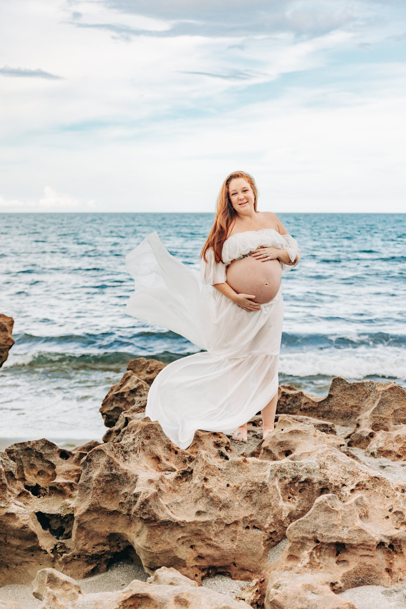 Maternity photographer Jupiter FL portrait of an expecting mother in a flowing white dress on the rocks at Coral Cove Beach.
