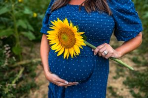 Pregnant woman in blue polka dot dress holding a bright yellow sunflower while cradling her baby bump outdoors.