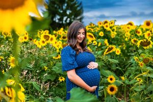 Pregnant woman in blue polka dot dress cradling baby bump in vibrant sunflower field under dramatic sky.