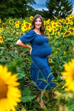 Pregnant woman in blue polka dot dress smiling in a vibrant sunflower field on a sunny day.