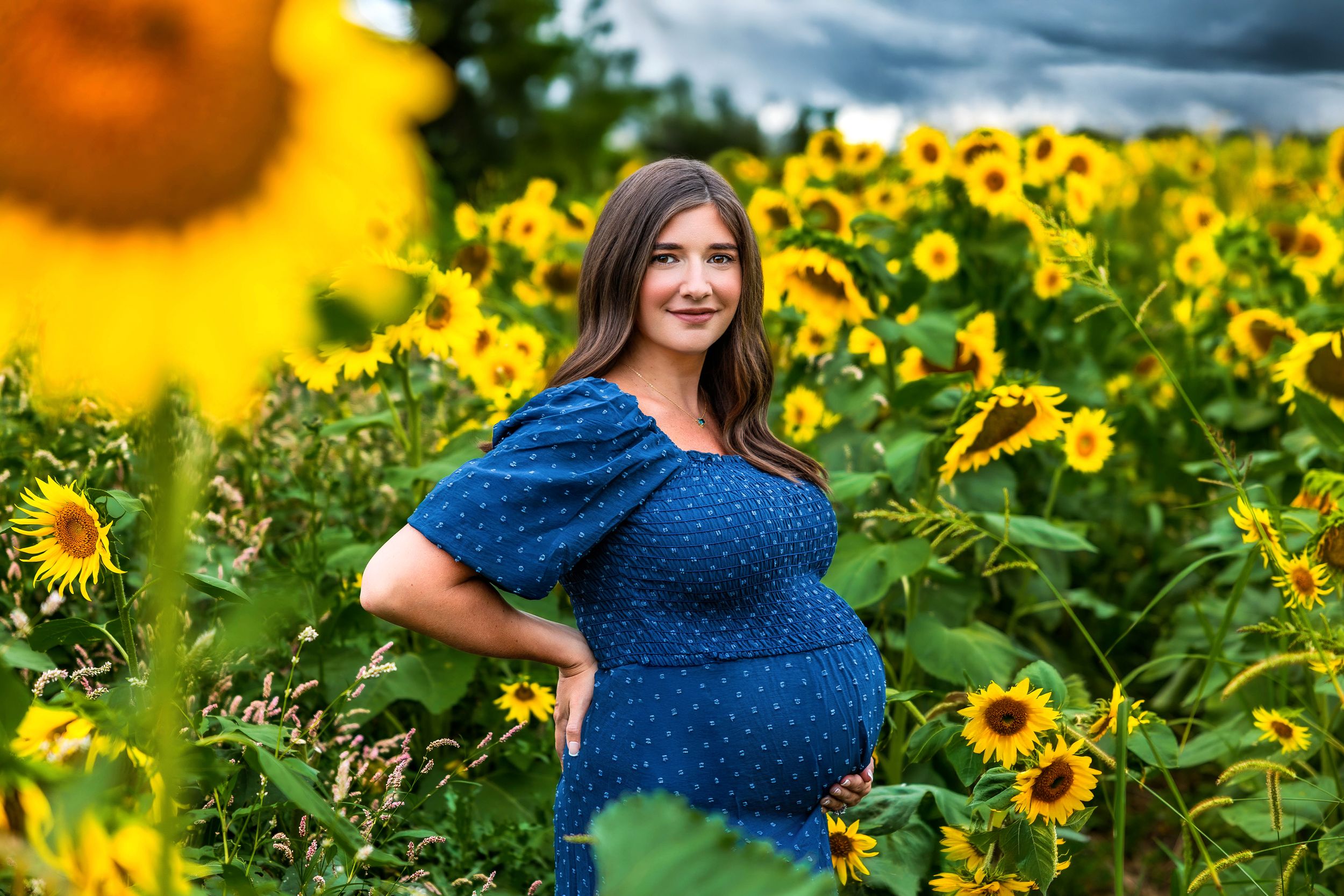 Pregnant woman in blue polka dot dress posing in vibrant sunflower field under dramatic cloudy sky.