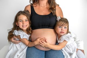 Pregnant woman in black crop top and jeans poses with two young girls in white dresses hugging her baby bump.