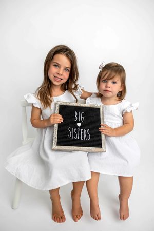 Two young girls in white dresses hold a black letter board sign reading Big Sisters against a white background.