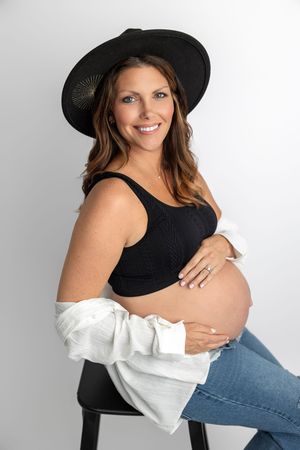 Smiling pregnant woman in black crop top, wide-brim hat, and jeans, cradling her baby bump while seated.