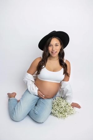 Pregnant woman in white crop top, jeans, and black hat sits on floor holding baby bump with white flowers nearby.