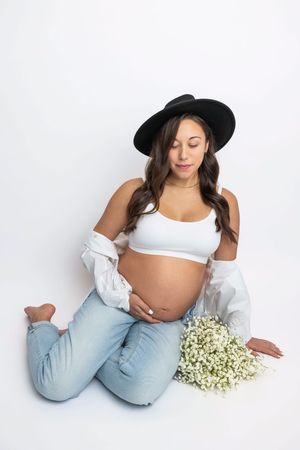 Pregnant woman in white crop top, jeans, and black hat sits on floor holding baby bump with white flowers nearby.