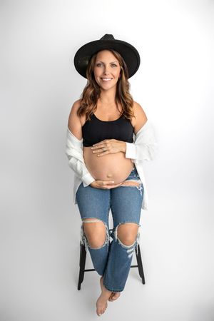 Smiling pregnant woman in black crop top, ripped jeans, and wide-brim hat sits on stool cradling baby bump.