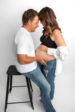 A couple shares a tender moment, hands on pregnant belly, foreheads touching, in a bright studio setting.