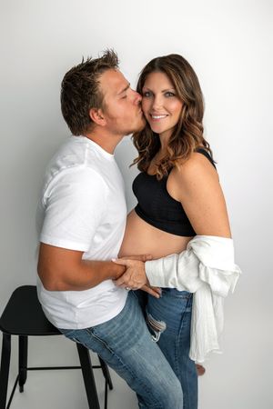 A man kisses a smiling pregnant woman on the cheek while holding her baby bump in a bright studio setting.