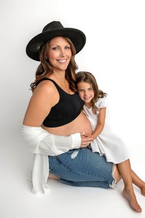 Pregnant woman in black crop top and hat sitting with young daughter hugging her baby bump on white background.