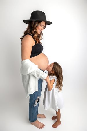 Pregnant woman in black crop top, jeans, and hat, with young girl in white dress kissing her baby bump.