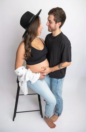 Pregnant couple posing together, woman in black crop top, hat, and jeans, man in black t-shirt holding her baby bump.