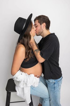 Pregnant couple sharing a tender kiss, woman in black hat and crop top with exposed baby bump, man in black shirt.