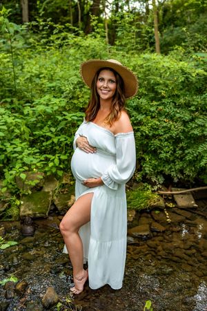 Pregnant woman in white off-shoulder dress and straw hat standing by a creek in lush green forest.