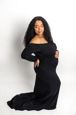 Pregnant woman in elegant black off-shoulder gown kneeling gracefully, cradling baby bump during maternity photoshoot.