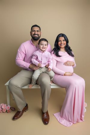 A happy family in matching pink outfits poses for a maternity photo, with a pregnant mother, father, and young son on a bench.