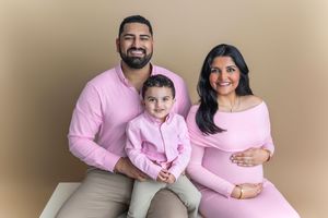 A smiling family of three in matching pink outfits, with a pregnant mother, father, and toddler posing for a maternity photo.