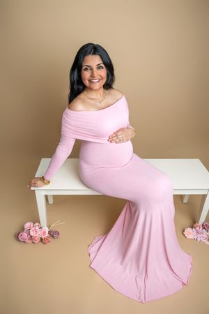 Pregnant woman in pink off-shoulder maternity gown sits on white bench with roses, smiling at camera.