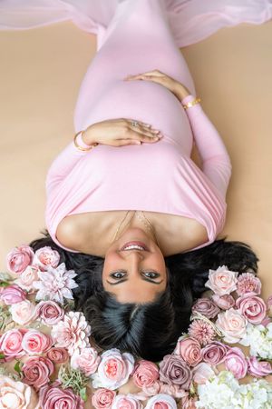 Pregnant woman in pink dress lying among pink roses and flowers, smiling upward in a beautiful maternity photo shoot.