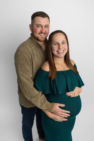 Smiling couple posing for maternity photo, woman in teal off-shoulder dress cradling baby bump, man in olive sweater embracing her.