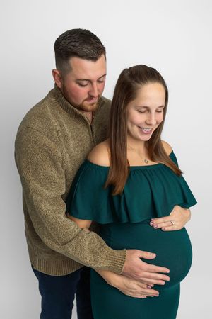 A couple shares a tender maternity moment, man embracing pregnant woman in teal dress against white background.