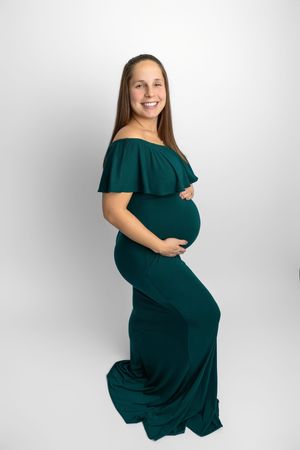 Smiling pregnant woman in elegant emerald green off-shoulder maternity gown, cradling her baby bump against white background.