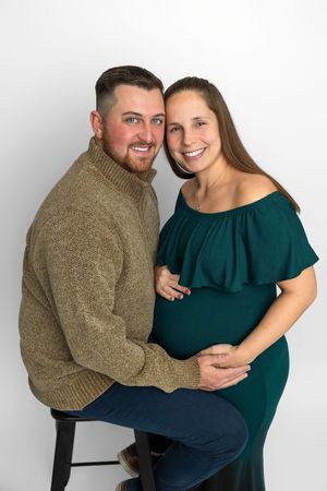 Smiling couple posing for maternity photo, man in olive sweater embracing pregnant woman in teal off-shoulder dress.