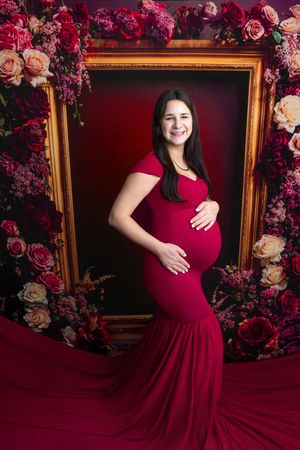 Pregnant woman in red mermaid gown poses by floral frame backdrop for elegant maternity portrait session.