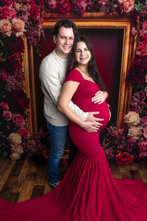 Couple poses for maternity photo shoot, woman in red gown cradling baby bump, floral backdrop with gold frame.