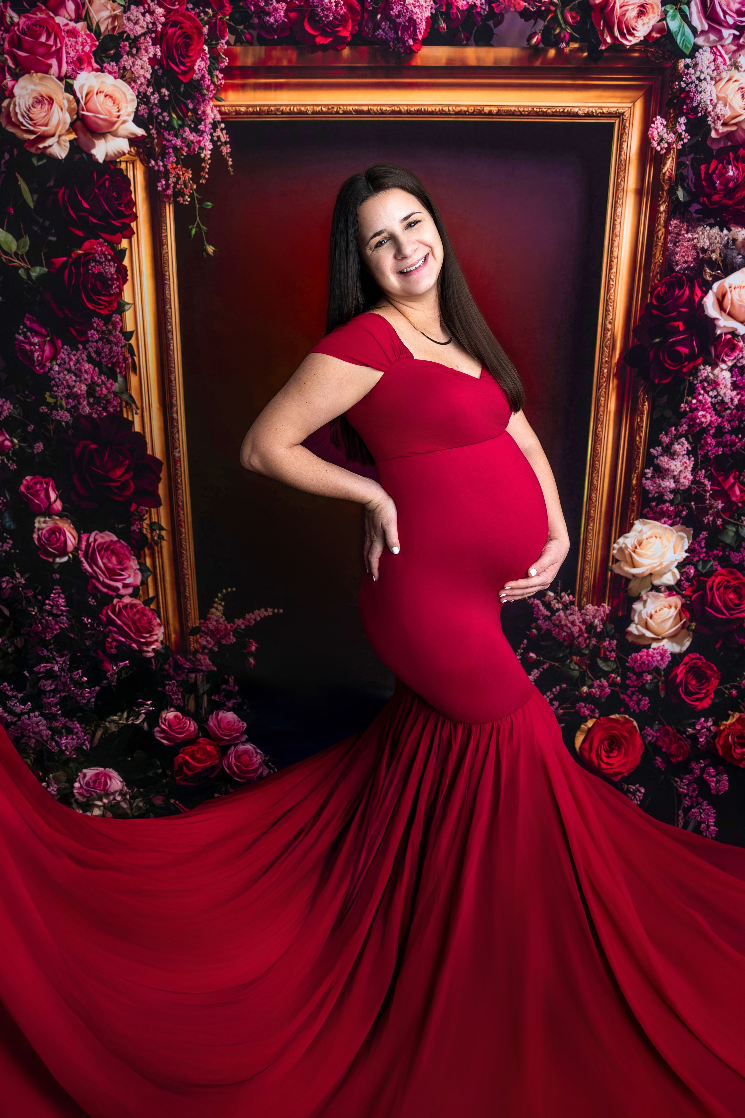 Pregnant woman in red mermaid gown poses among flowers before a gold frame backdrop.