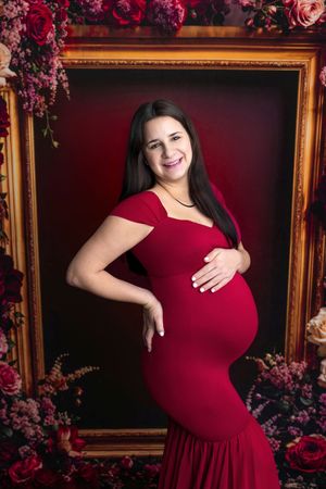 Smiling pregnant woman in red maternity gown poses in front of floral-framed backdrop.