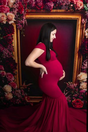 Pregnant woman in red mermaid gown cradling baby bump, posed before floral frame backdrop.