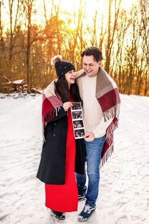 Happy couple sharing ultrasound photos outdoors in snowy winter forest at golden sunset, wrapped in plaid blanket scarf.