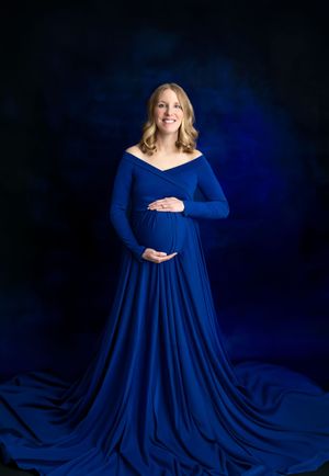 Pregnant woman in elegant royal blue off-shoulder maternity gown with long train poses against dark blue backdrop.