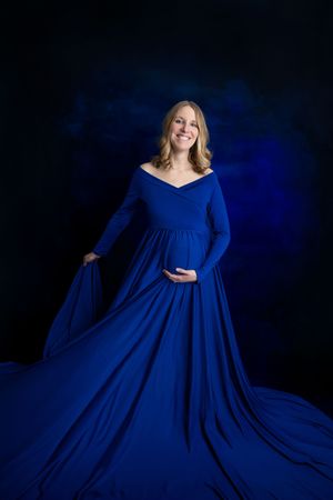 Smiling pregnant woman in elegant royal blue off-shoulder gown with flowing train poses against dark studio backdrop.