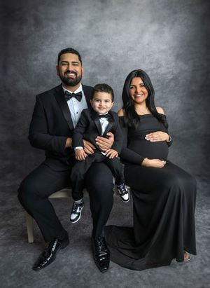 Family maternity photo with parents and toddler in matching black formal wear against gray studio backdrop.