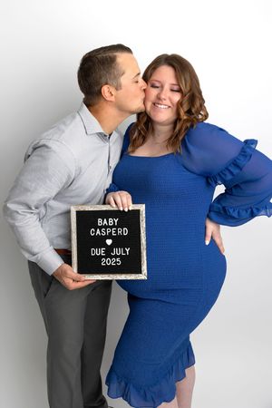 Pregnant woman in blue dress holds baby announcement sign with partner kissing her cheek, due July 2025.