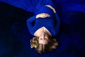 Pregnant woman in blue dress lying on blue fabric, cradling baby bump, with flowing blonde hair spread around her.