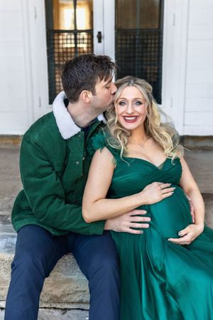 Couple in matching green outfits sharing a tender moment, pregnant woman smiling as man kisses her cheek outdoors.