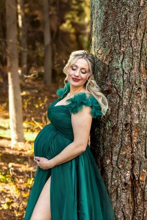 Pregnant woman in elegant green tulle gown leaning against tree in forest, cradling baby bump, blonde hair, outdoor maternity photo.