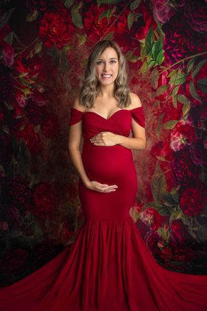 Pregnant woman in red off-shoulder mermaid gown poses against floral backdrop for maternity photo session.