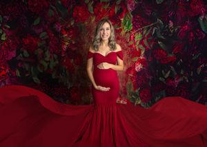 Pregnant woman in red off-shoulder mermaid gown poses against floral backdrop for maternity photo.