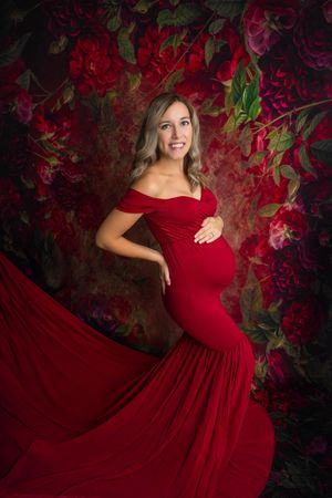 Pregnant woman in elegant red off-shoulder mermaid gown poses against a floral backdrop for a maternity portrait.