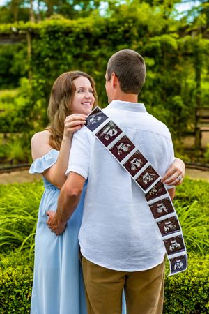Pregnant couple shares ultrasound strip in lush garden, woman smiles as man holds sonogram photos behind his back.