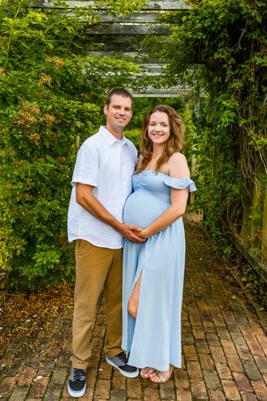 Smiling couple poses in green garden tunnel, pregnant woman in blue dress with partner in white shirt holding her baby bump.