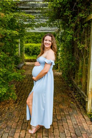 Smiling pregnant woman in a light blue off-shoulder maxi dress stands on a brick path surrounded by lush green garden pergola.