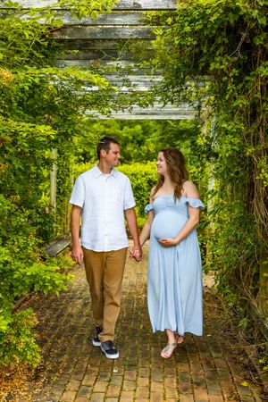 Pregnant couple holding hands and walking through a lush green garden pergola, woman in blue dress cradling baby bump.