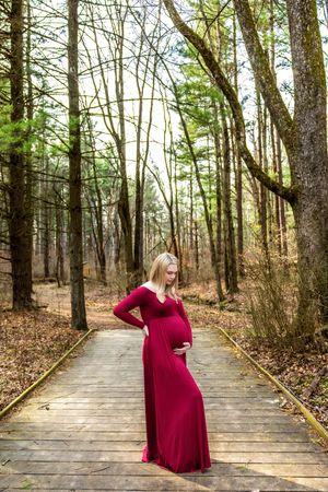 Pregnant woman in red maxi dress standing on wooden boardwalk path in forest, cradling baby bump.