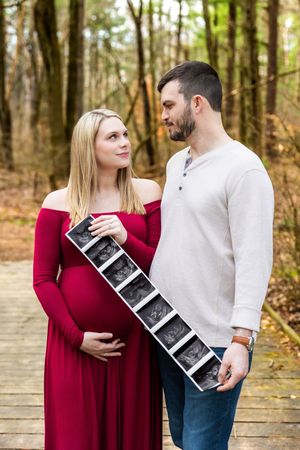 Pregnant woman in red dress and man hold ultrasound photos on a woodland boardwalk during maternity photoshoot.